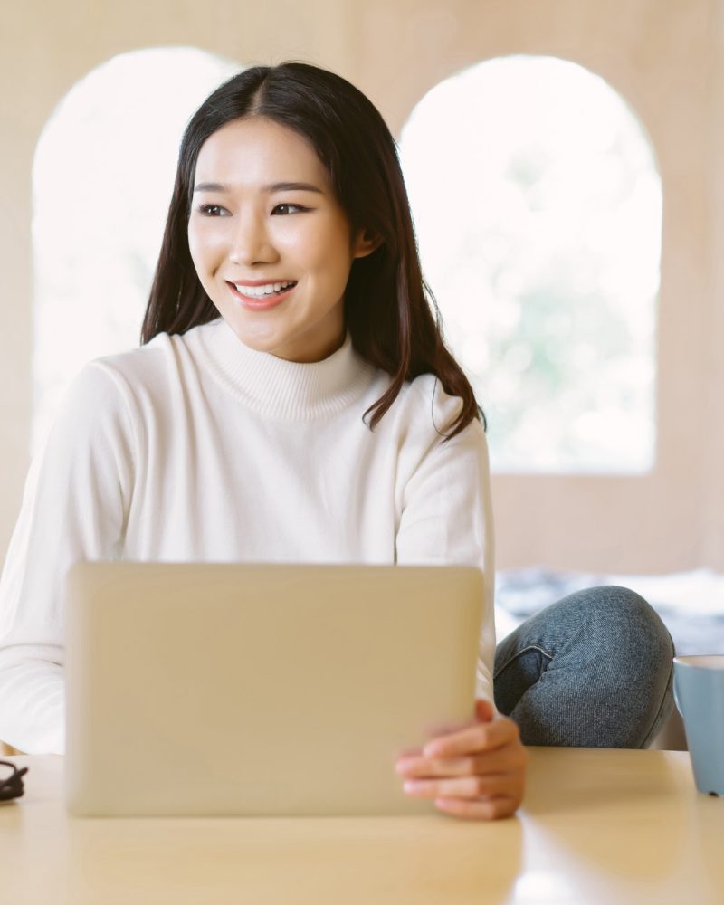 smiling freelancer Asian woman work remotely on laptop and looking away in home winter atmosphere.